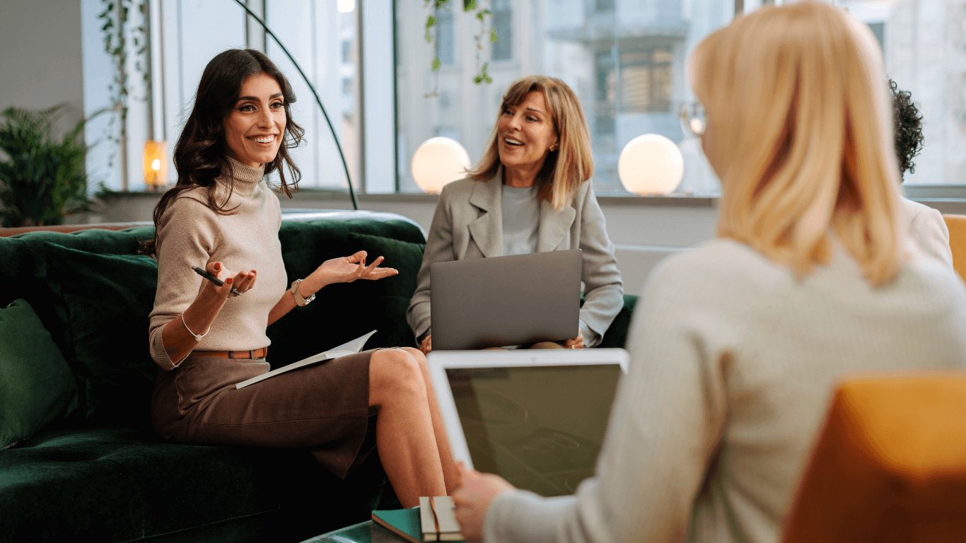 Three woman professionals sitting on a green velvet couch with laptops while smiling.